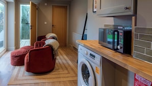 A compact living space with a washing machine, a microwave, and a fire blanket on the counter. There are red bean bags and a wooden door. - 4 Bed Holiday Home by Guestz (Rhosneigr)