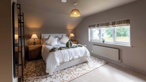 A bedroom with a bed, two bedside lamps, a window with curtains, a radiator, and a patterned rug. - 4 Bed Holiday Home by Guestz (Rhosneigr)