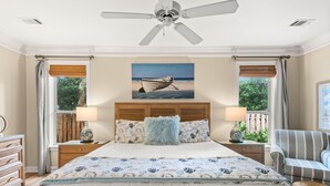 Une chambre à coucher avec un lit, deux tables de chevet, un ventilateur de plafond, une fenêtre avec des rideaux, un tableau représentant un bateau et une chaise.