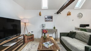 Una sala de estar moderna con un televisor de pantalla plana, una mesa de centro de madera y un sofá verde. Hay dos ventanas con tragaluces, un escritorio con lámpara y un reloj en la pared.