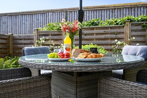 Une table ronde en verre, entourée de chaises en osier, une bouteille de jus de fruits, une assiette de fruits, une miche de pain et une tasse de café.