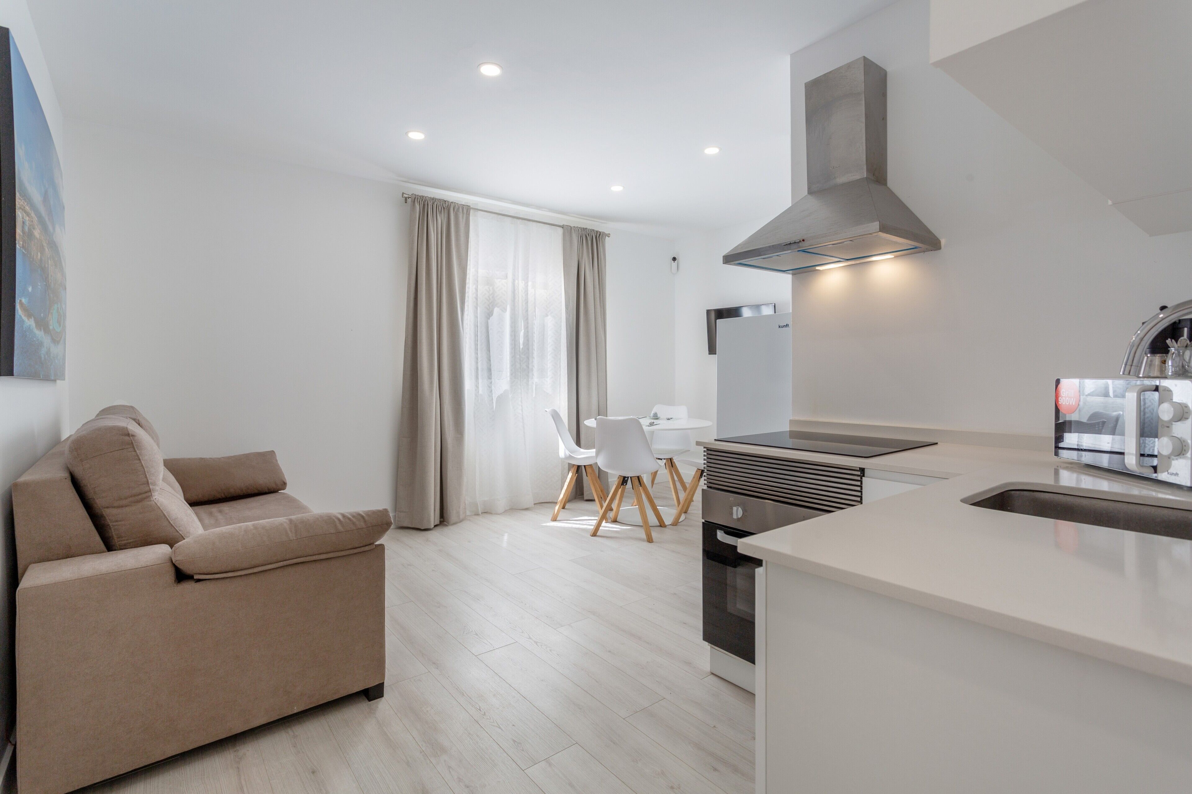 Une cuisine moderne dotée d’un four encastré, un coin repas avec une table et des chaises blanches, et un canapé beige.