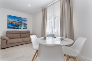 A modern room with a round dining table, white chairs, a beige sofa, and a large window with curtains.