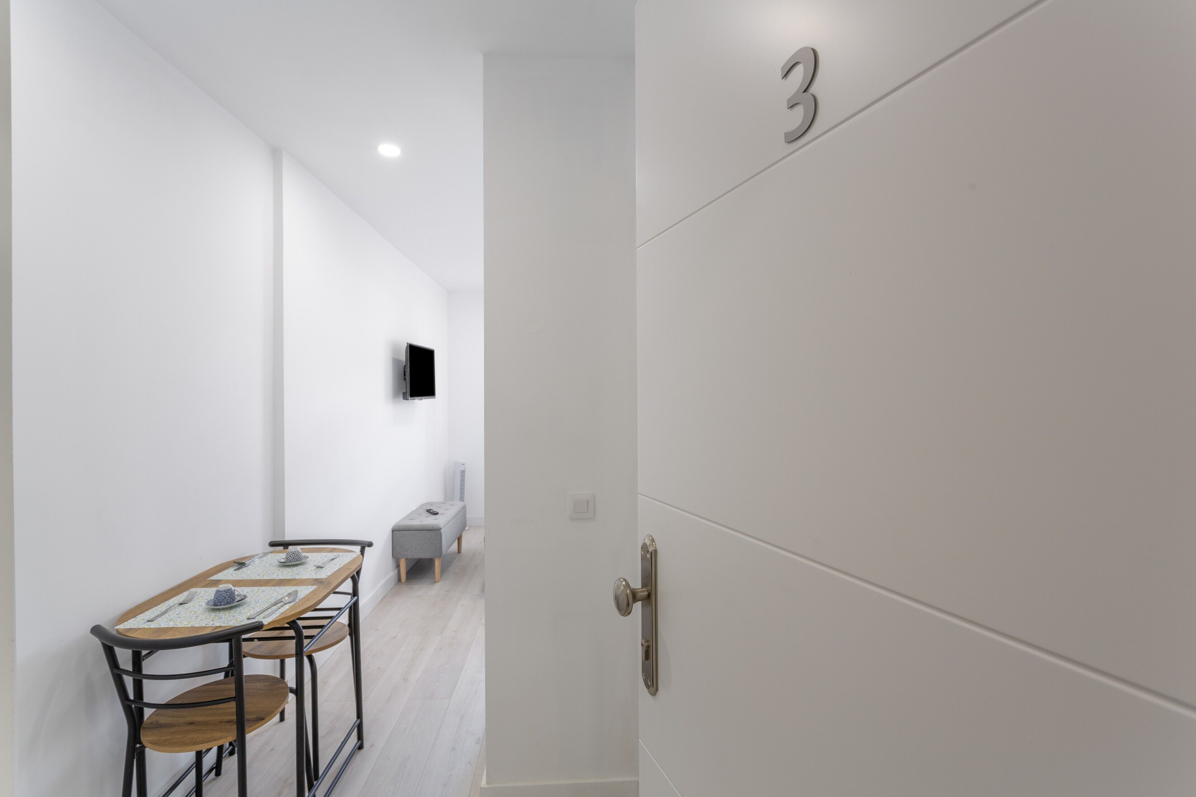 Ein modernes Zimmer mit Esstisch, Stühlen, einer Bank, einem an der Wand befestigten Fernseher und einer Tür mit einer Nummer.