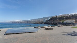 A coastal promenade with stone flooring, a pool, and buildings in the background.