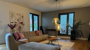 Una sala moderna con un sofá seccional, un reposapiés y una pequeña mesa de centro. Hay dos ventanas amplias con persianas, una lámpara de pie y un cuadro abstracto de gran tamaño en la pared.
