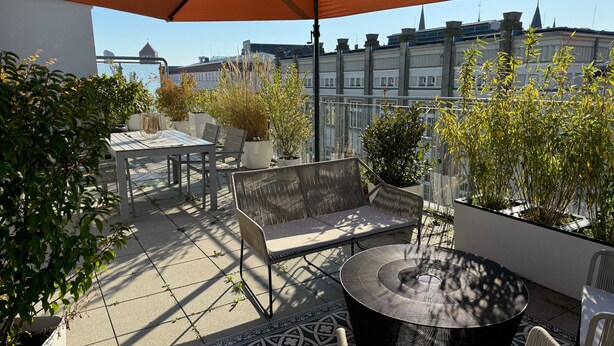 Un patio en la azotea con mobiliario exterior, plantas en macetas y vistas a edificios al fondo.