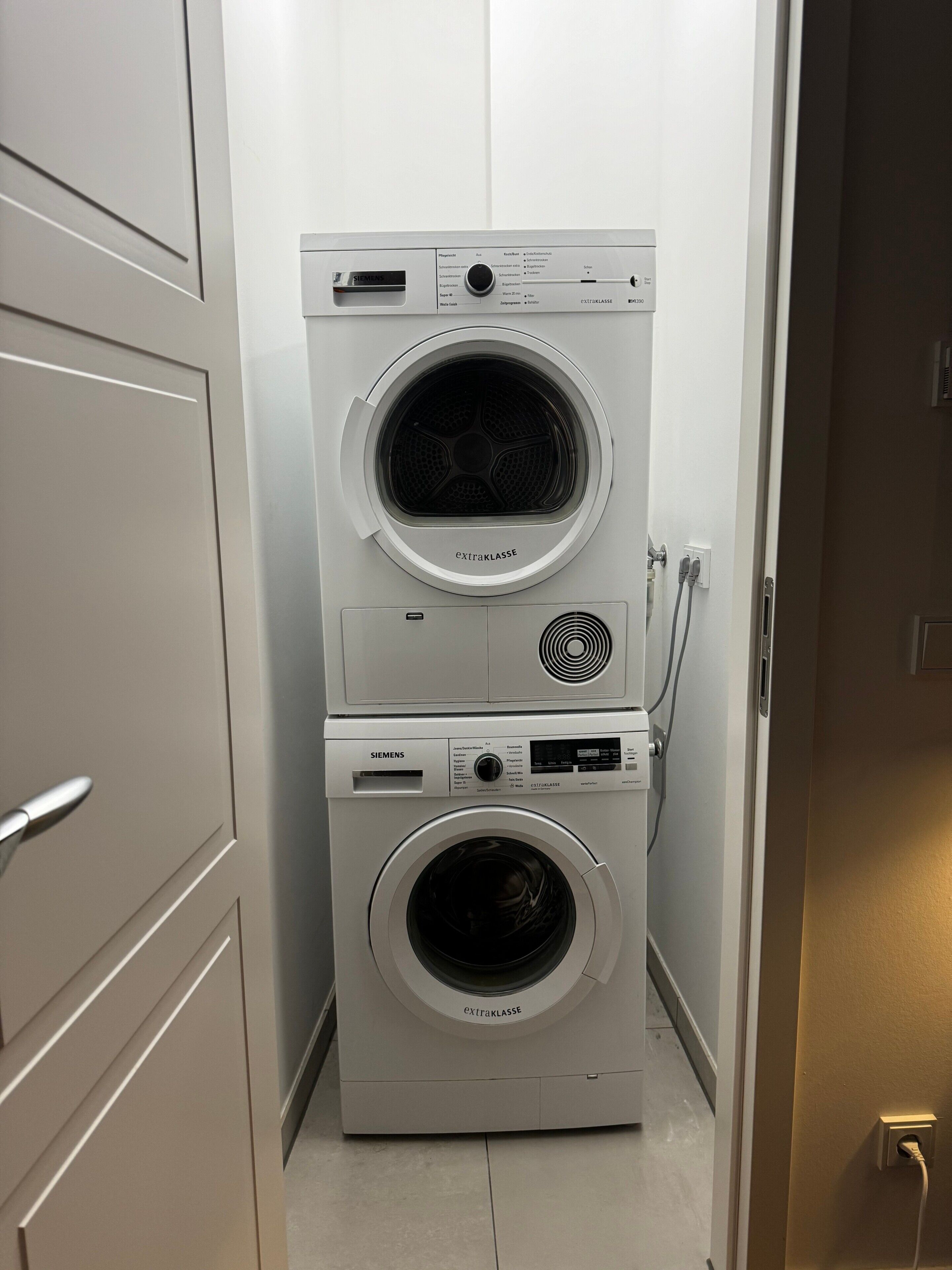 A laundry room with two stacked washing machines, a door, and a wall socket.