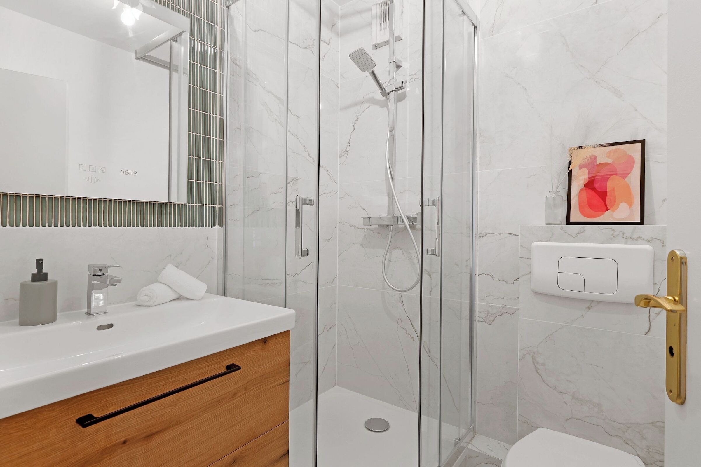 Un bany modern amb plat de dutxa envoltat de vidre, un lavabo blanc amb un armari de fusta i un quadre abstracte emmarcat a la paret.