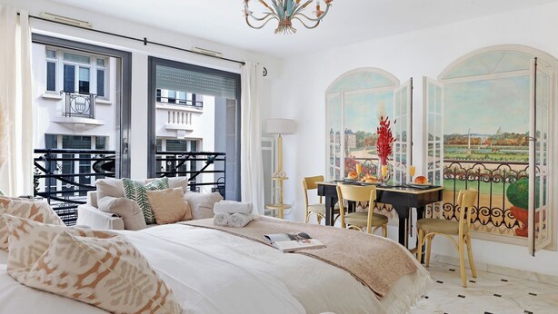 A hotel room with a large bed, a dining table with chairs, a balcony with a railing, and a view of a cityscape through the windows.