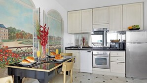 A kitchen with white cabinets, a dining table with chairs, a painting on the wall, and a view of a park.