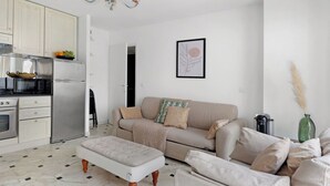 A modern living room with a sofa, ottoman, and a dining table. There is a kitchen area with a refrigerator, microwave, and cabinets. A chandelier hangs from the ceiling, and a framed artwork is on the wall.