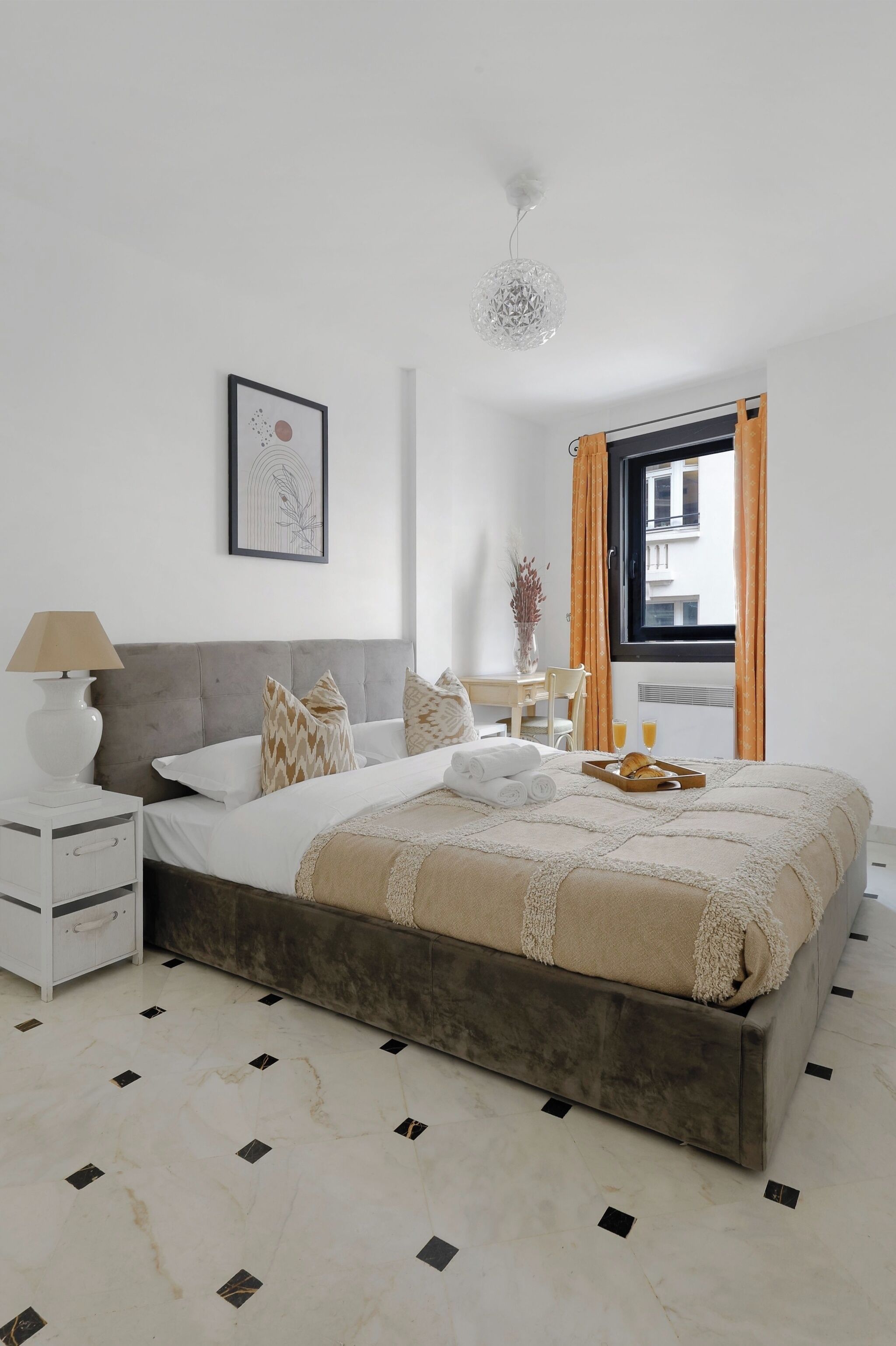 A modern bedroom with a large bed, a bedside table, a lamp, a framed picture on the wall, and a window with curtains.