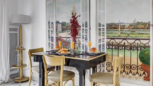 A dining area with a table set for two, a vase with red flowers, and a view of a cityscape through open windows.