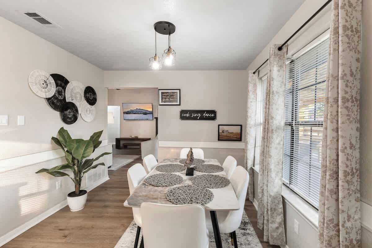 Een eethoek met een tafel, stoelen, een potplant, wanddecoratie en een raam met gordijnen.