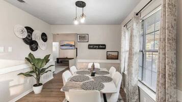 Een eethoek met een tafel, stoelen, een potplant, wanddecoratie en een raam met gordijnen.