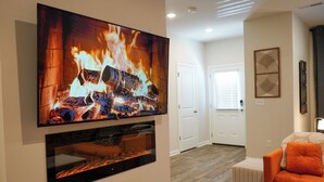 Una sala de estar con una televisión de pantalla plana grande montada en la pared mostrando una escena de fuego, una chimenea debajo, un sofá naranja y una puerta que da a otra habitación.