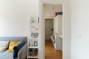 Una habitación moderna y bien iluminada, con un sofá gris, un estante blanco con diversos objetos, una lavadora y una puerta que da a un baño.