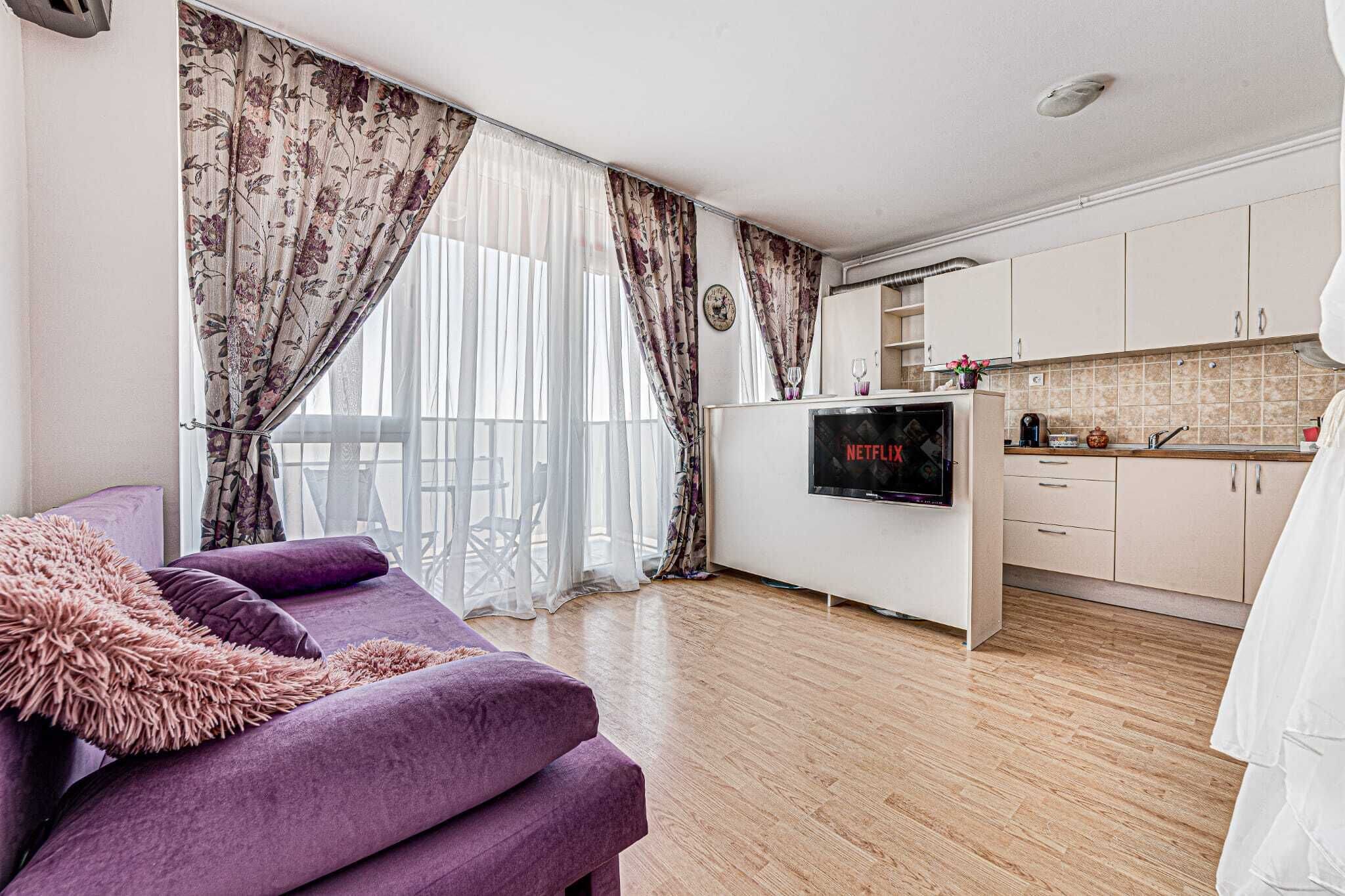 A living room with a purple sofa, a flat-screen TV, and a kitchen area with cabinets and a microwave.