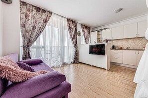 A living room with a purple sofa, a flat-screen TV, and a kitchen area with cabinets and a microwave.