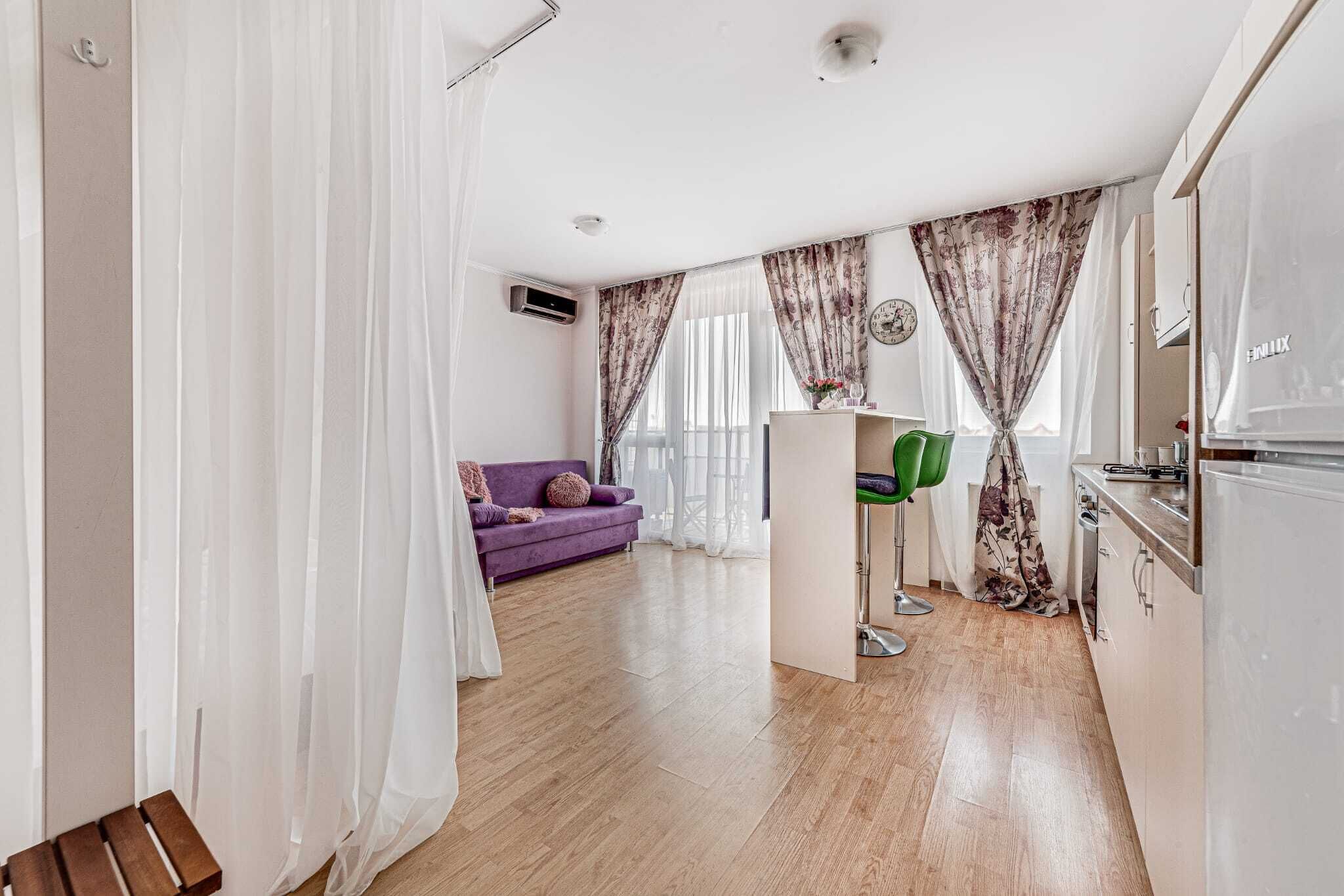 Ein modernes Wohnzimmer mit einem violetten Sofa, einem grünen Sessel und einer Küchenzeile inklusive Kühlschrank.
