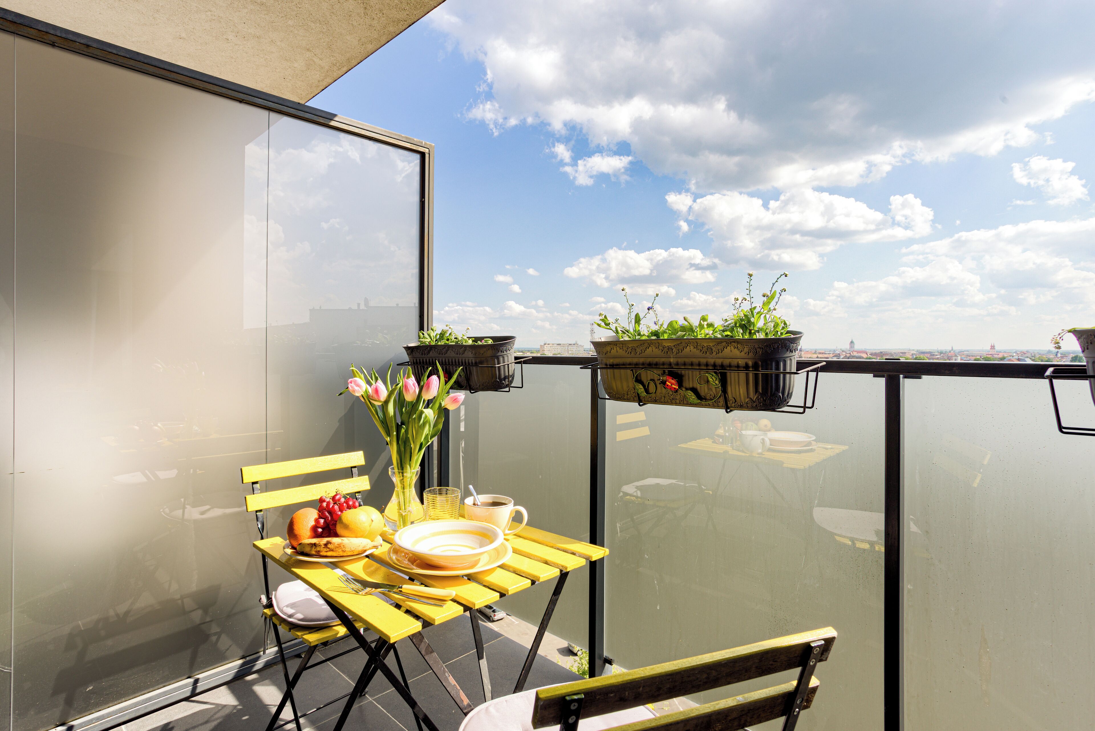 A balcony with a yellow table set for breakfast, a vase with flowers, and potted plants.