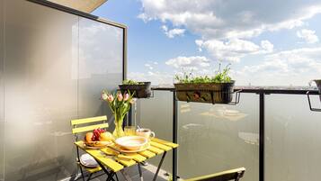 A balcony with a yellow table set for breakfast, a vase with flowers, and potted plants.