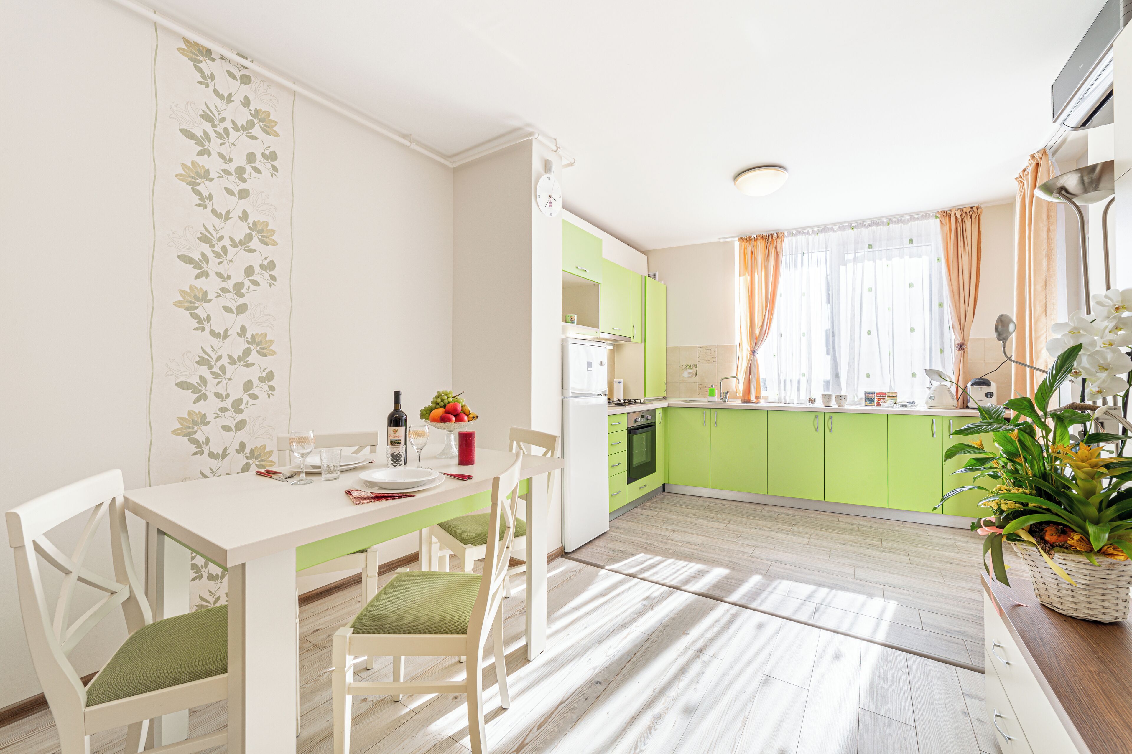 Une cuisine moderne avec des meubles de couleur verte, une table à manger blanche avec des chaises et une plante en pot posée sur le comptoir.