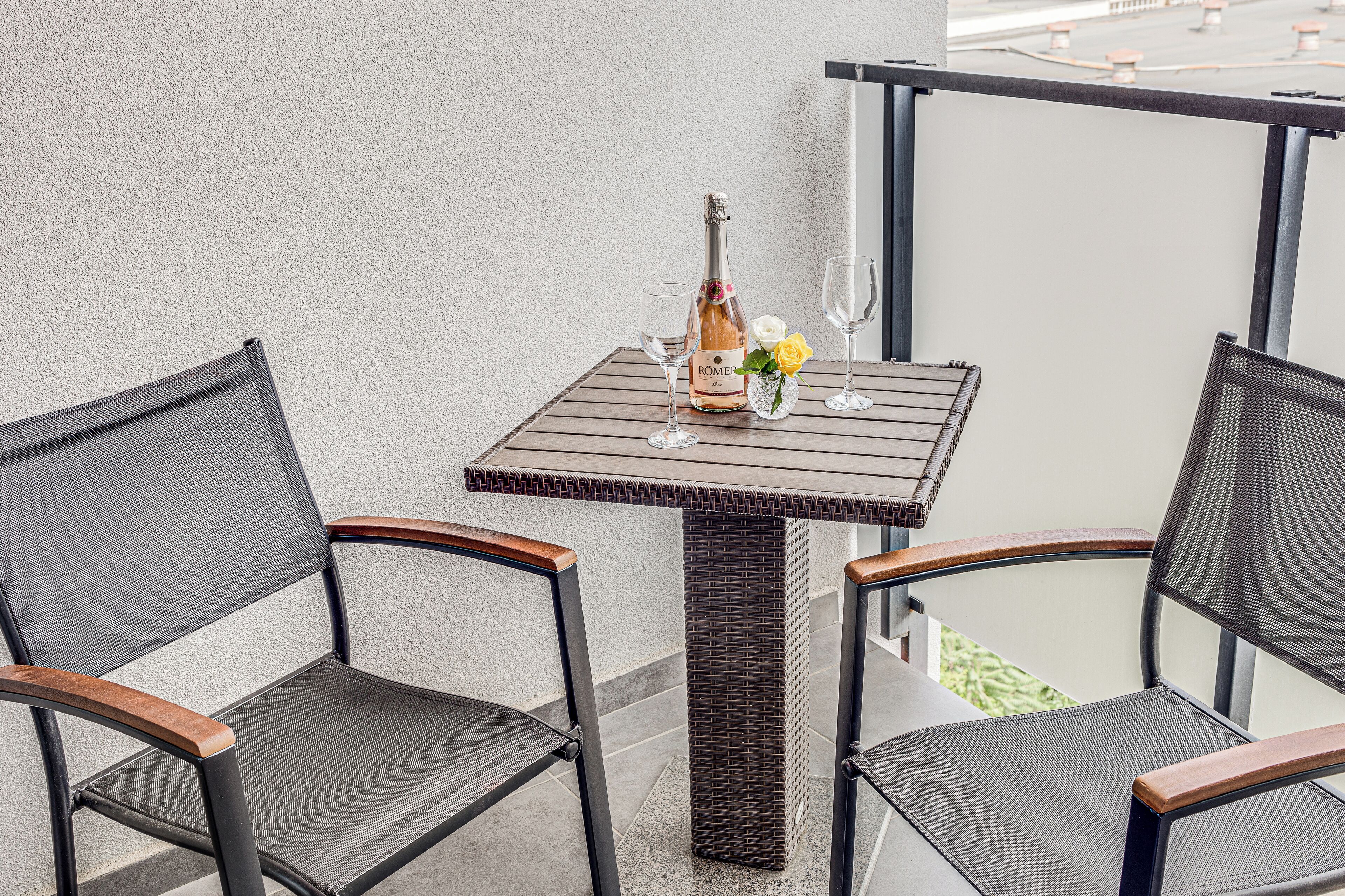 A small outdoor balcony with a square table, two chairs, a bottle of wine, and a vase with flowers.