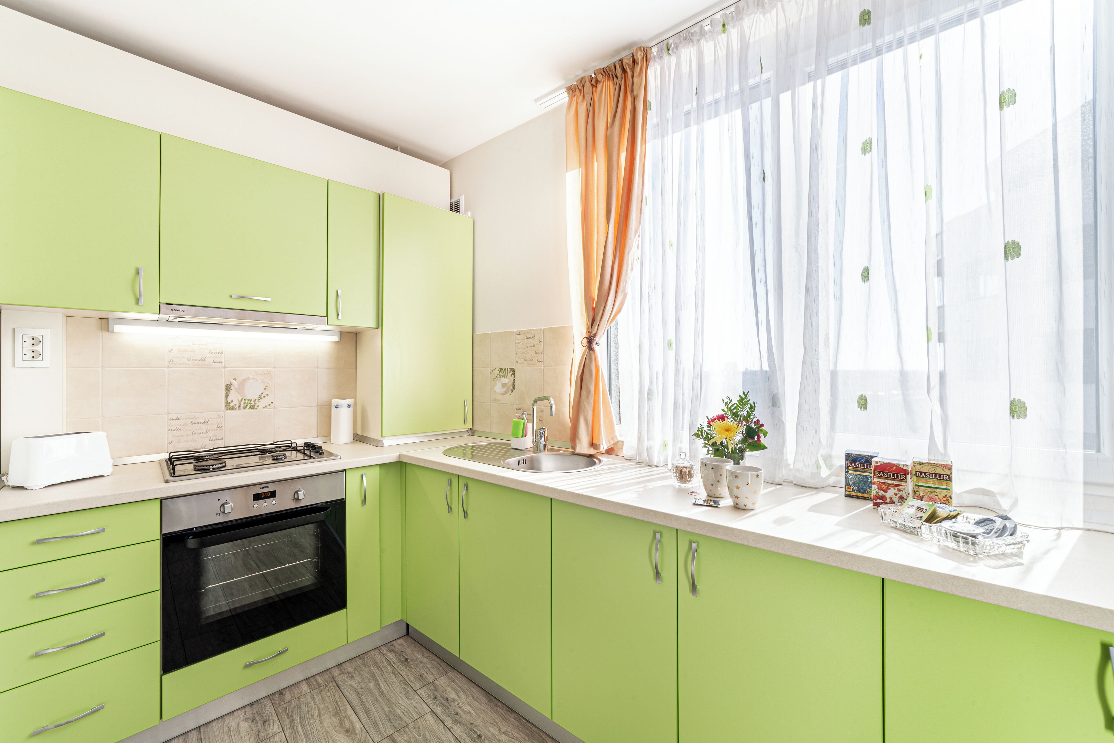 Una cocina con armarios verdes, una estufa y una ventana con cortinas translúcidas.