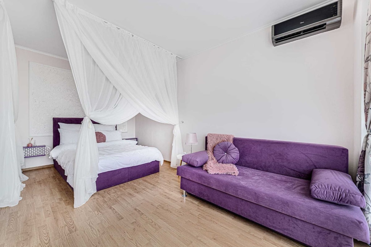 Ein Schlafzimmer mit einem Himmelbett, einem violetten Sofa und einem Nachttisch mit Lampe.
