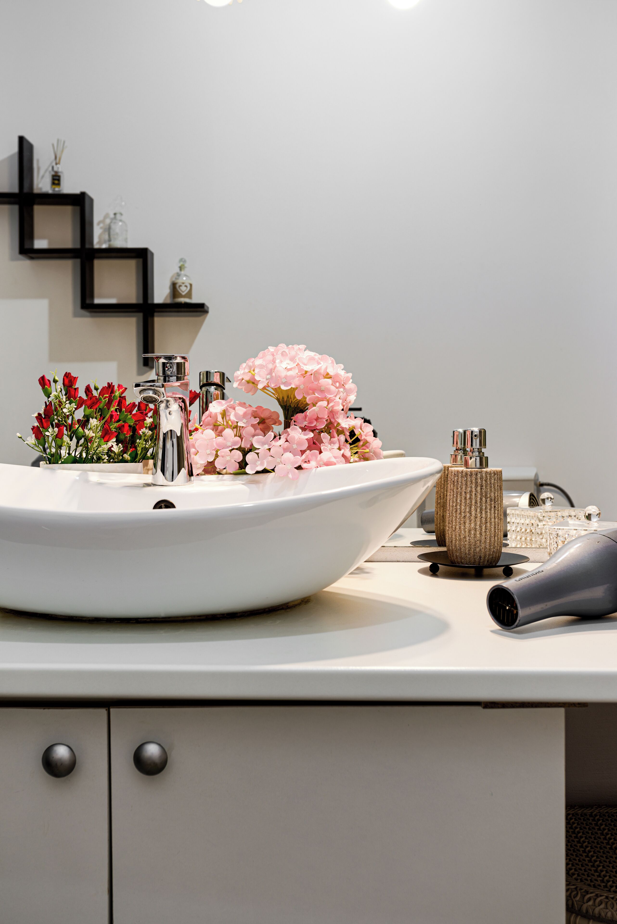 A bathroom with a large white sink, a mirror, a black shelf with decorative items, and a vase with pink flowers.