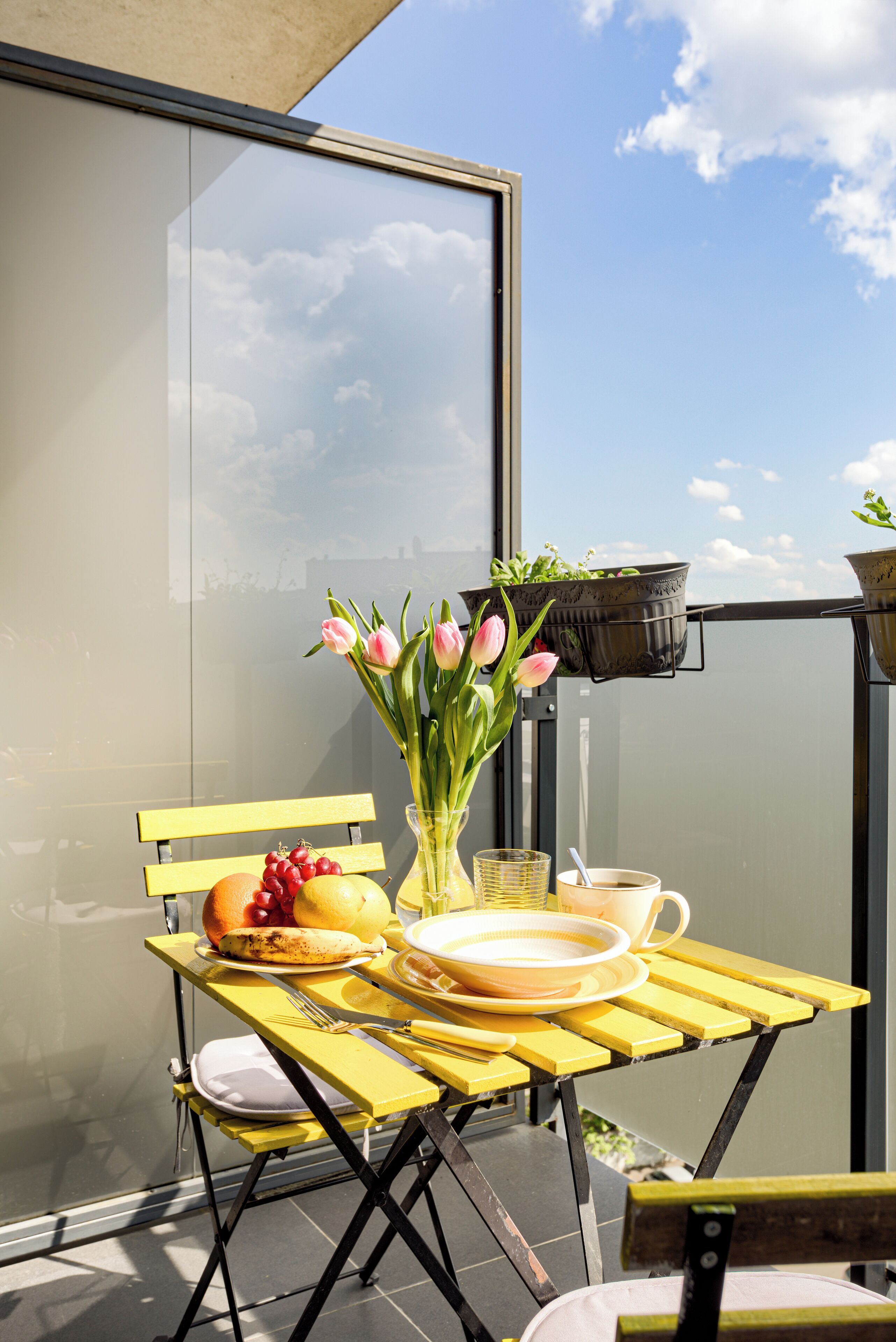 A yellow table with a vase of pink tulips, a bowl of fruit, and a plate of food on a balcony with a view of the sky and clouds.