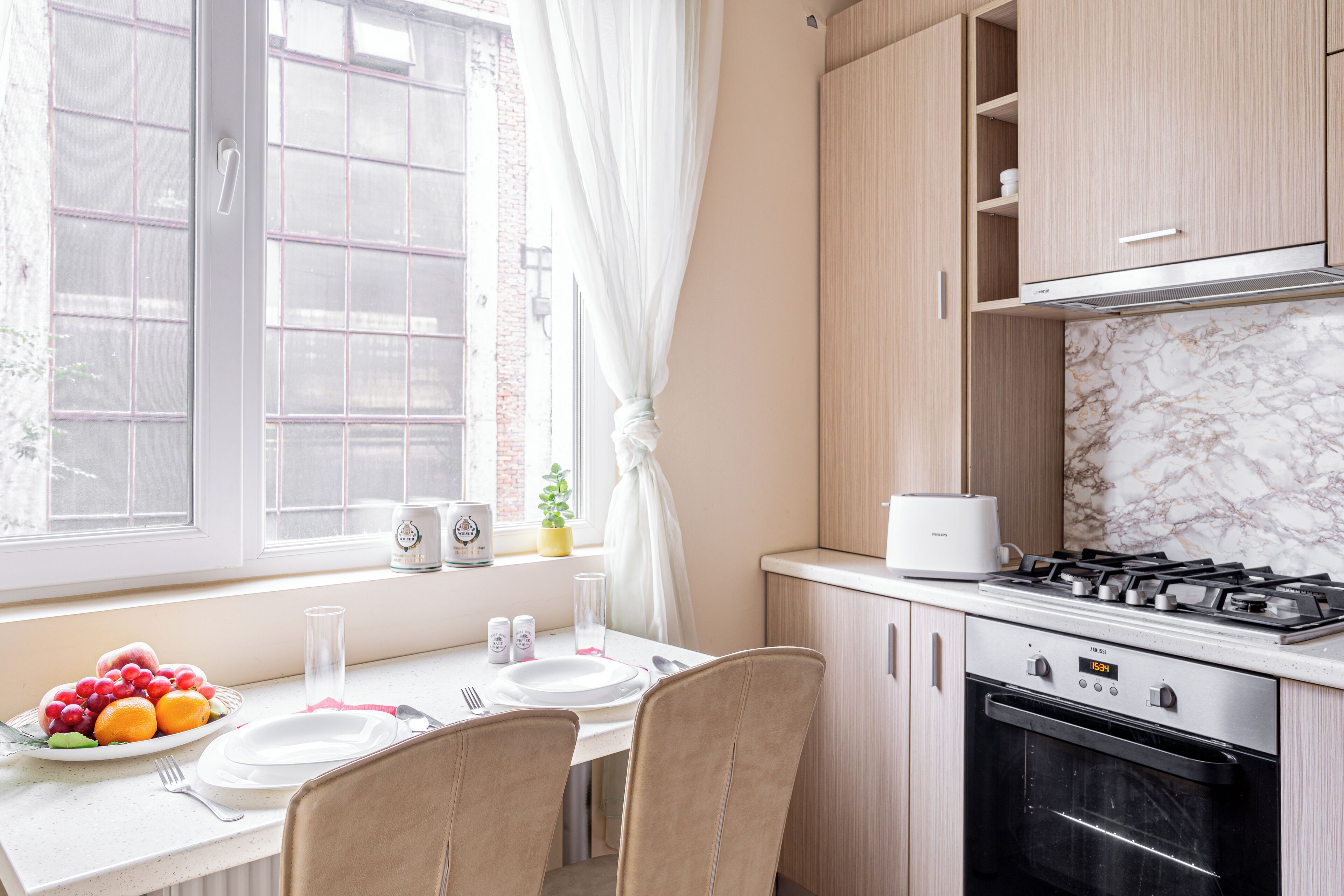 Une cuisine avec une table à manger pour deux personnes, un fourneau et une fenêtre avec des rideaux.