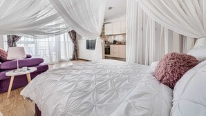A spacious bedroom with a large bed, a small seating area, and a view of the kitchen. - ARI - ARED Luxurious Apartments (Arad)