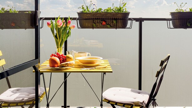 A balcony with a table set for two, a chair, and two hanging planters with flowers. - ARI - ARED Luxurious Apartments (Arad)