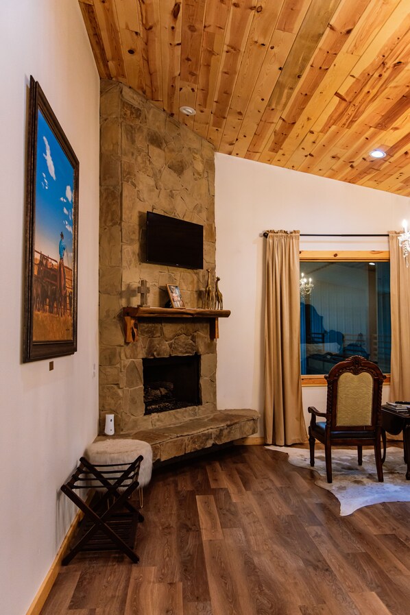 A cozy living room with a stone fireplace, wooden ceiling, and a large window with curtains.