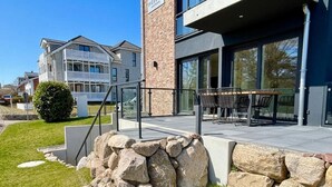 Un bâtiment moderne avec une façade en pierre, un espace extérieur aménagé avec une zone de sièges couverte et une pelouse bien entretenue.