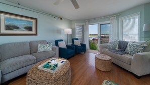 Una sala de estar con un sofá gris, sillones azules, un reposapiés de mimbre y un ventilador de techo.