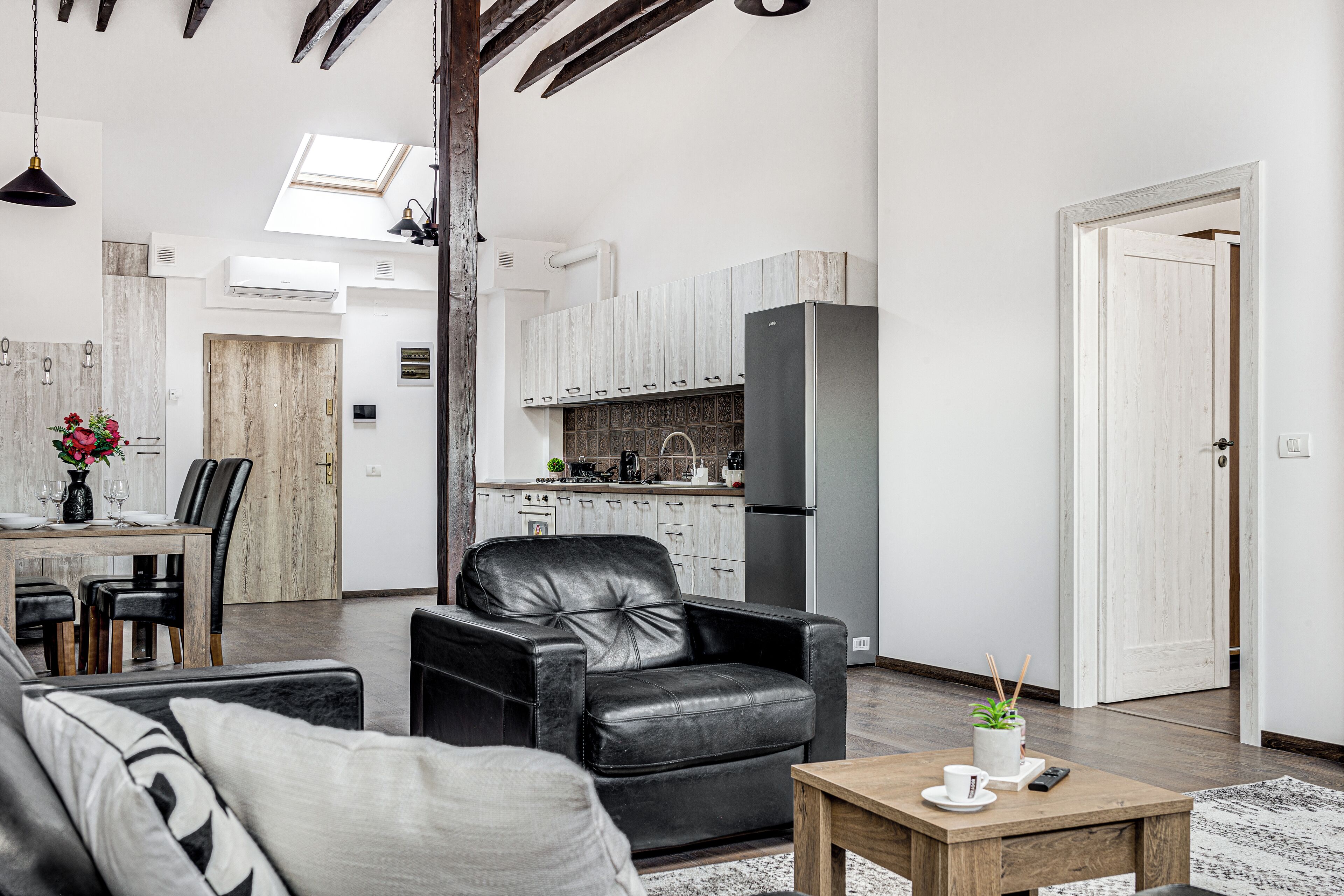 Una sala de estar moderna con un sofá de cuero, una mesa de centro de madera, y un comedor con una mesa y sillas de madera. Se ve una cocina con armarios blancos y un refrigerador al fondo.