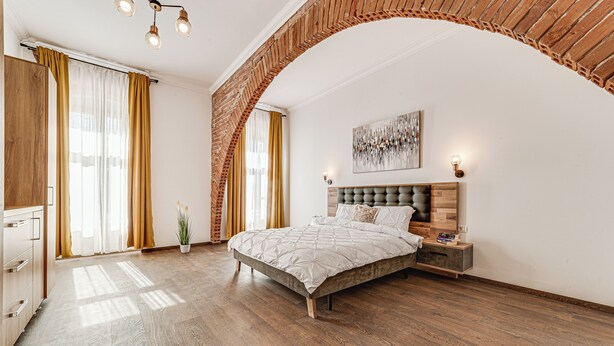 Une chambre dotée d’un grand lit, d’un plancher en bois, d’une arche en briques et d’un lustre moderne.