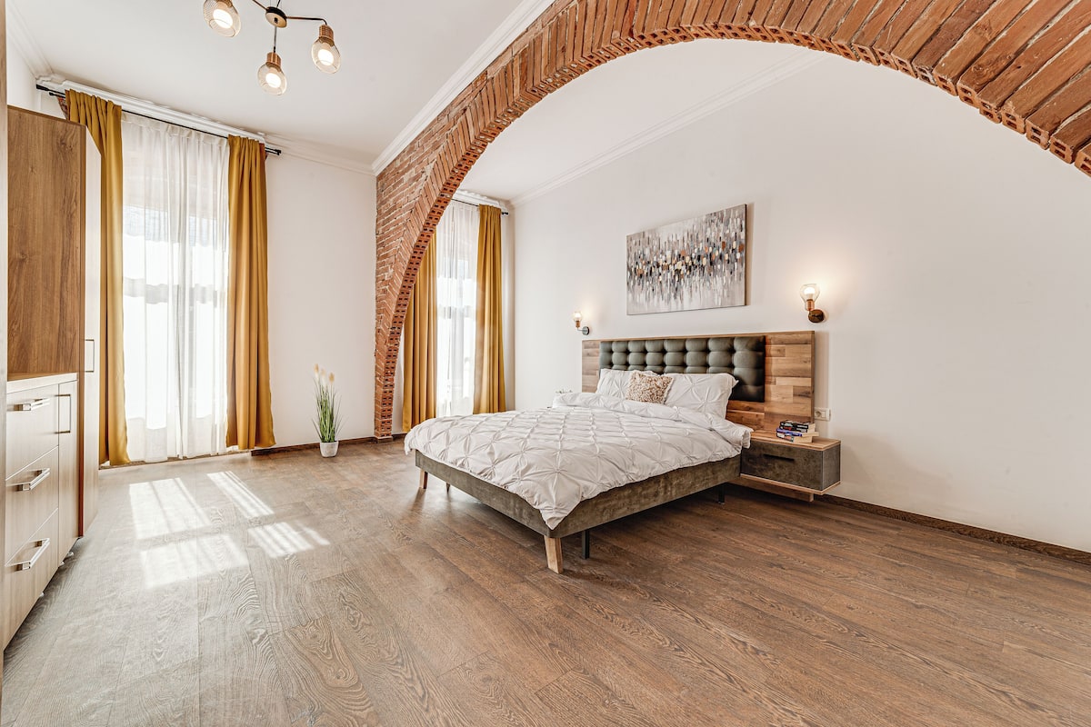 Une chambre dotée d’un grand lit, d’un plancher en bois, d’une arche en briques et d’un lustre moderne.