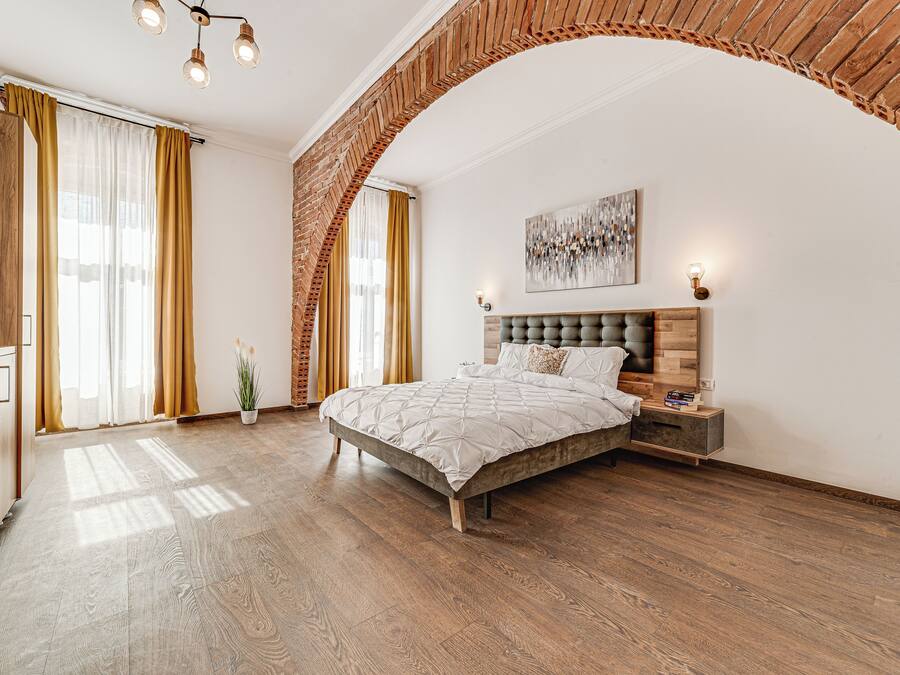 A bedroom with a large bed, wooden flooring, a brick archway, and a modern chandelier.