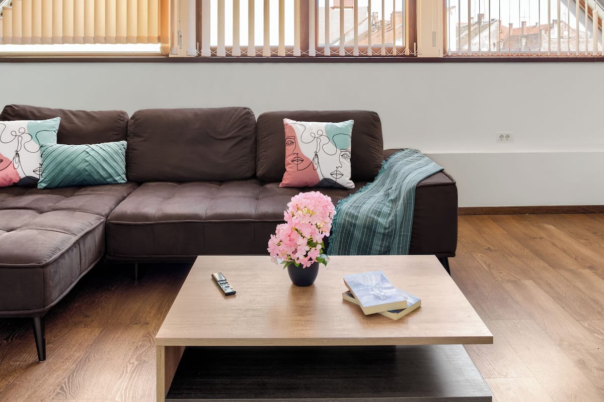 Un salon moderne doté d’un plafond en pente, d’un canapé d’angle marron, d’une table basse en bois et d’une fenêtre avec des stores verticaux.