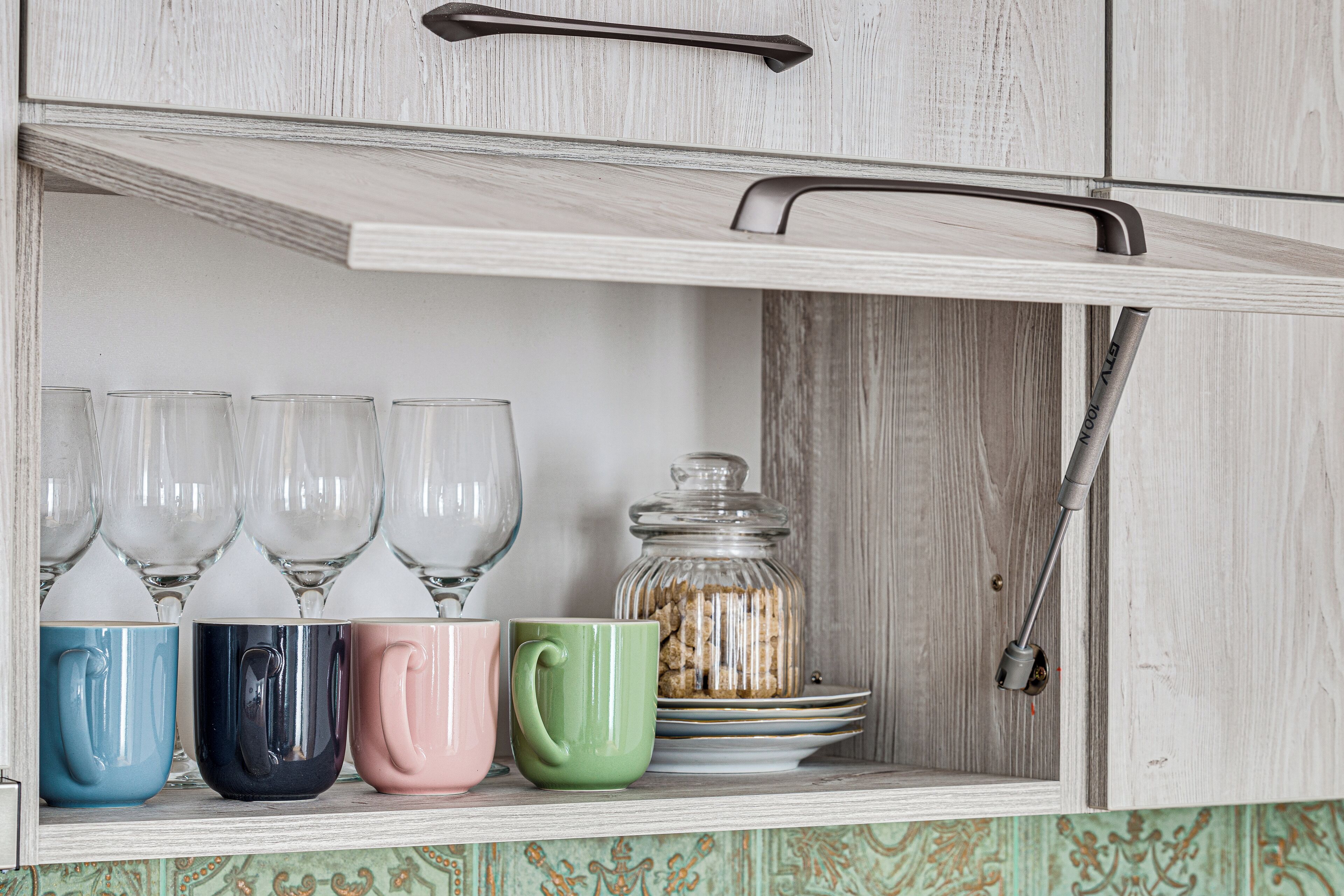 A kitchen cabinet with a handle, a shelf with clear glasses, and a glass jar with cookies.