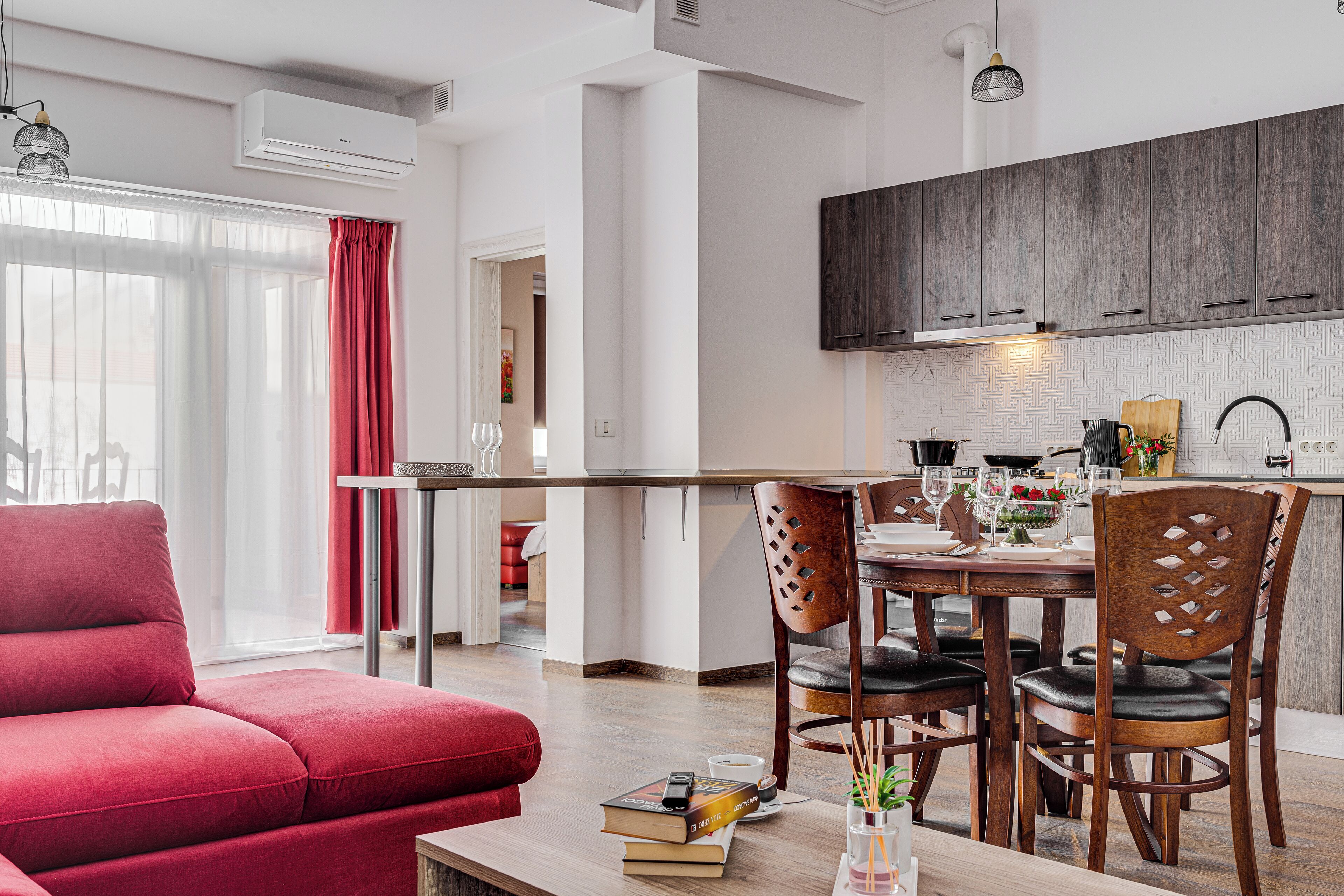 Una sala de estar moderna con un sofá rojo, un comedor con una mesa y sillas de madera, y una cocina con armarios de madera oscura.