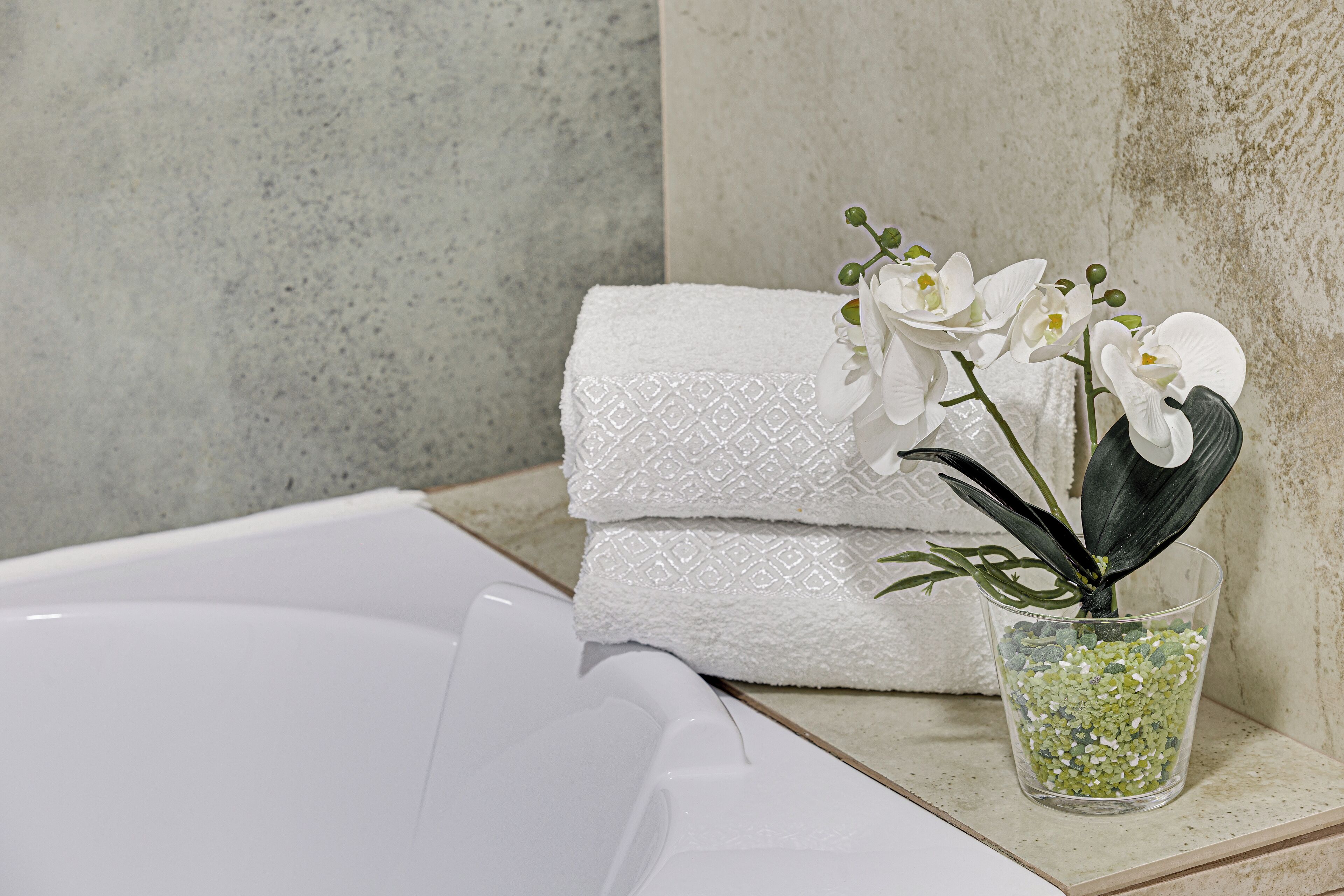 A bathroom with a white bathtub, a stack of white towels, and a glass vase with white flowers.