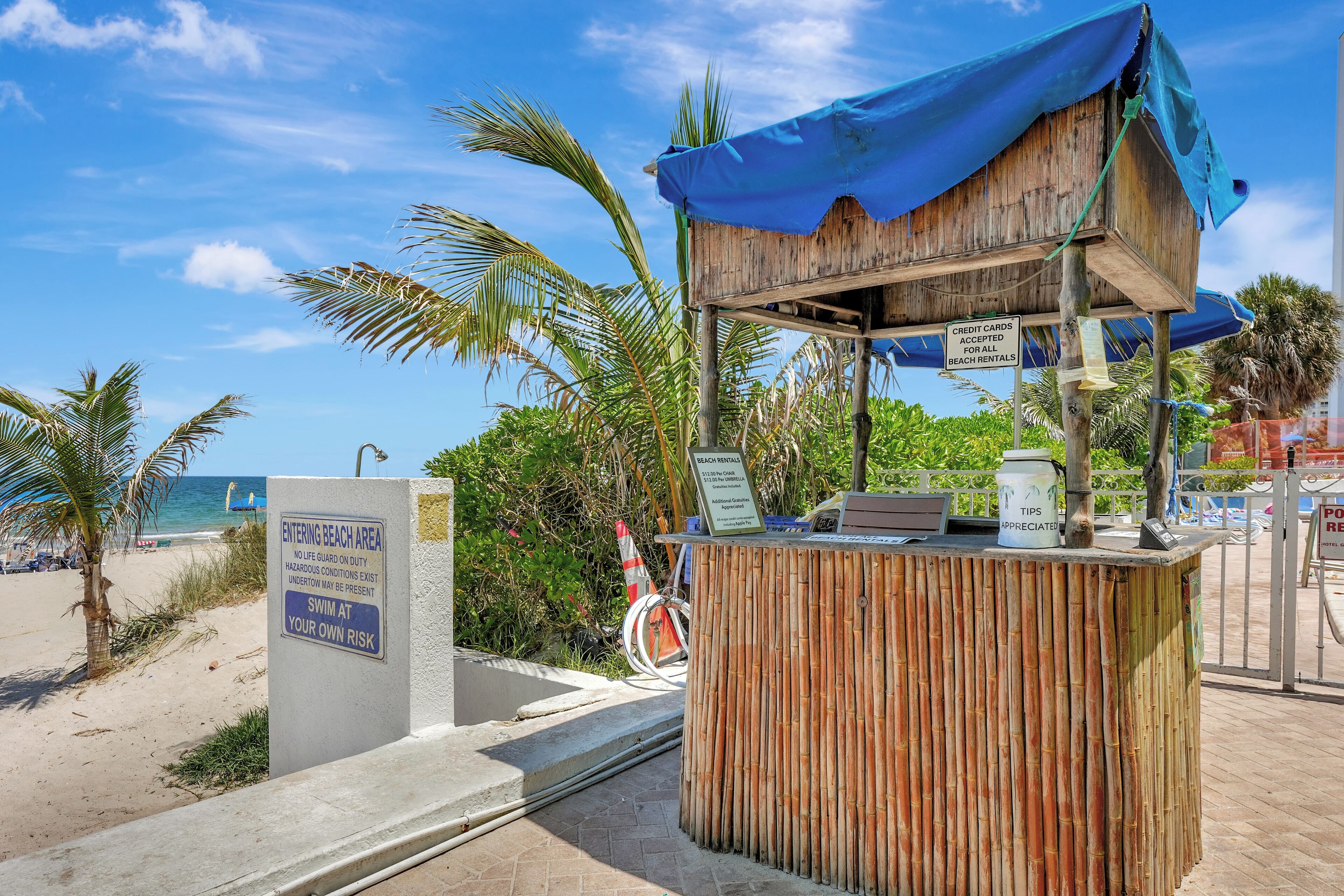 Un petit kiosque situé en bord de plage, avec un toit en toile bleue, une enseigne indiquant que les cartes de crédit sont acceptées, et une autre signalant les dangers potentiels.