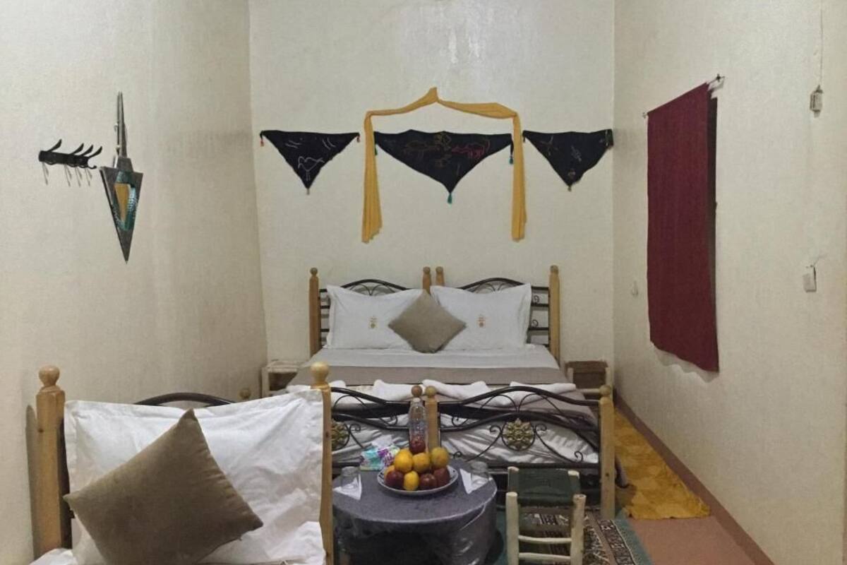 Ein Schlafzimmer mit einem Bett, einem Nachttisch mit Obstschale, einer Pendelleuchte und einem roten Vorhang.