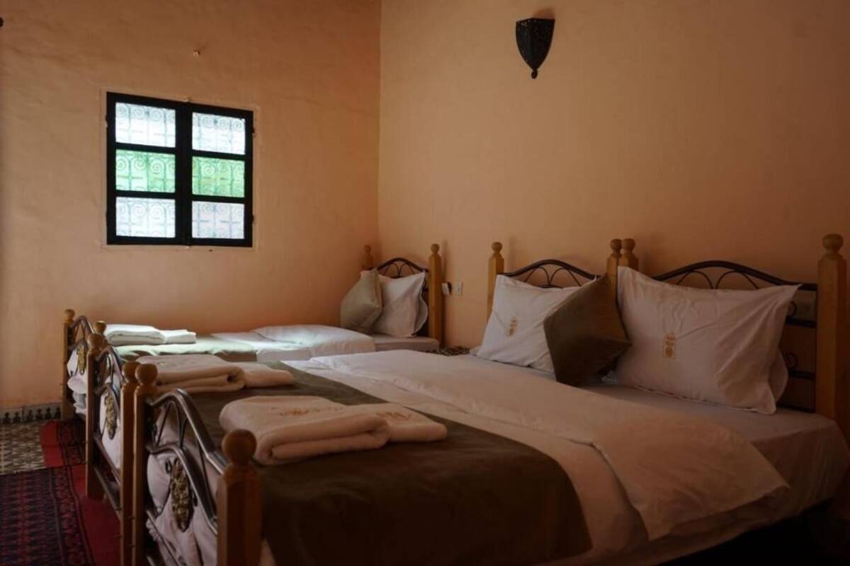 Ein Zimmer mit zwei Einzelbetten, jeweils mit Holz Kopfteilen und weißer Bettwäsche. Ein kleines Fenster mit dekorativem Muster, und eine Wandleuchte über den Betten.
